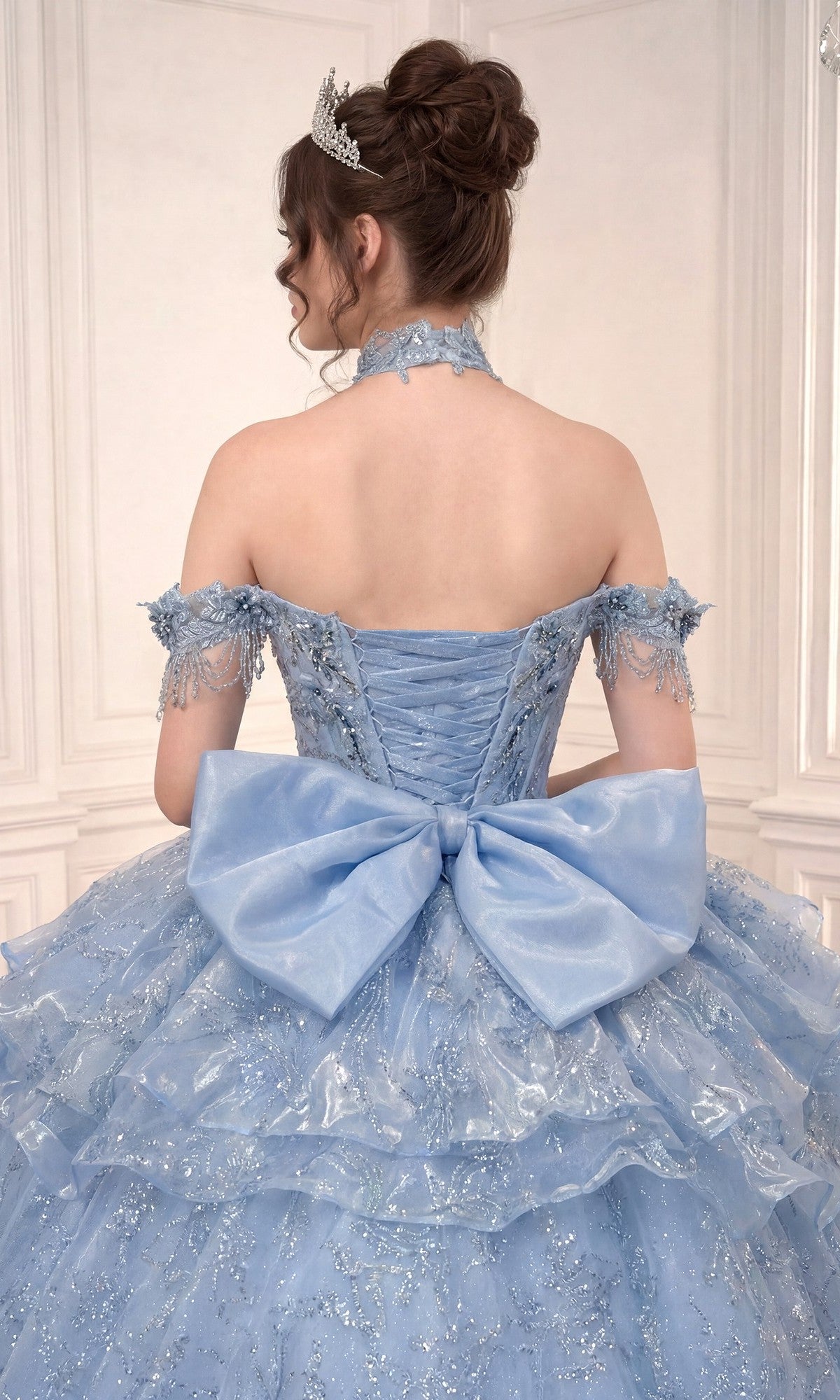 A woman in the Quinceanera Dress 15742 By Ladivine, a light blue off-the-shoulder gown with lace details, a large back bow, and an elegant detachable skirt, stands facing away in an ornate room.